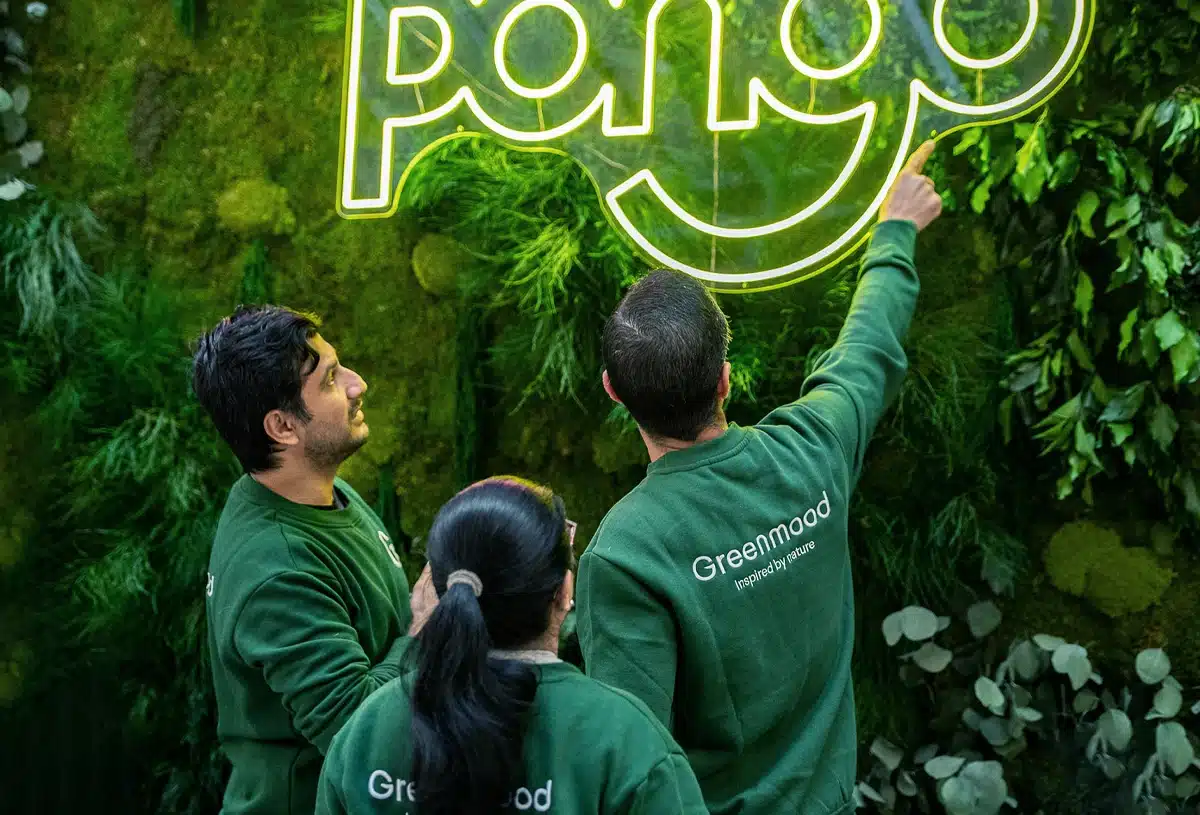 Three people admire illuminated sign amid greenery.